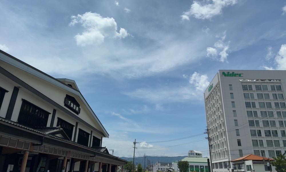 下諏訪駅前の風景。左に下諏訪駅、右にNidecの建物があり、青空と雲が広がっている。