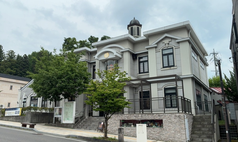 ニデックオルゴール記念館すわのねの外観。道路沿いに洋風の建物が建ち、入口付近に案内看板と木が見えている。